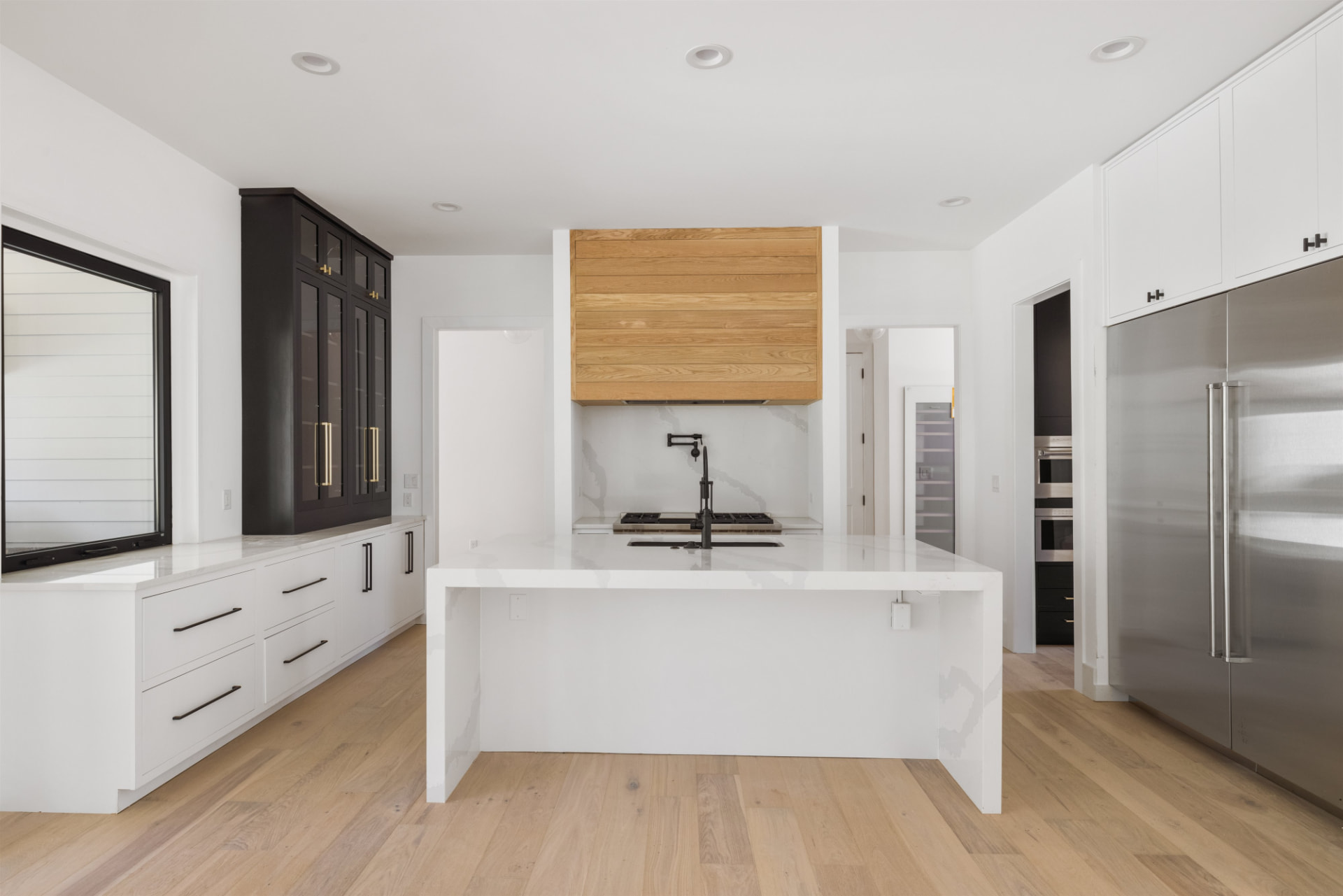 White flat-panel kitchen with waterfall island, oak range hood and black glass-front cabinets